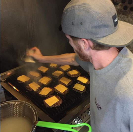 Man grilling square cheese slices on a barbecue, wearing a gray cap in a smoky outdoor cooking area