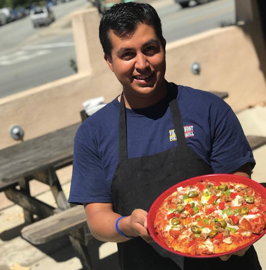 Person in a navy shirt holding a pizza on a sunny patio.