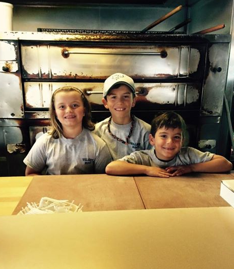 Three smiling children peeking over cardboard boxes in a workshop or warehouse setting