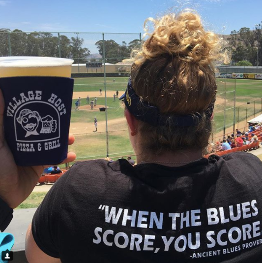 Fan at baseball game holding drink cup, wearing black shirt that says “WHEN THE BLUES SCORE, YOU SCORE”