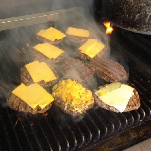 Cheeseburgers and sliced cheese cooking on a smoky grill, with a foil-wrapped item in the center
