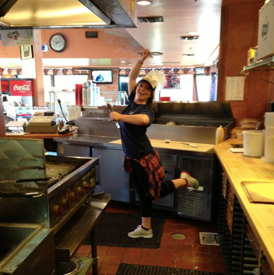 Person posing playfully in a diner kitchen, with a red plaid shirt tied at the waist
