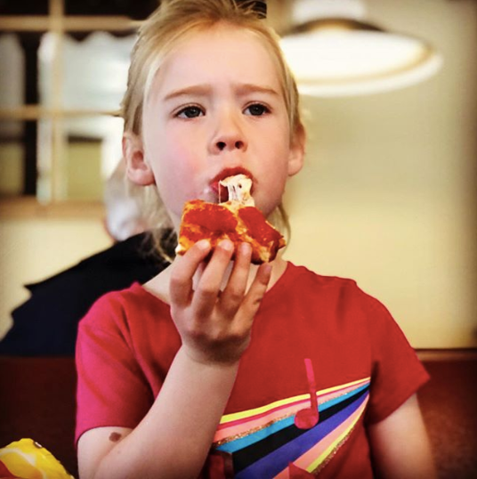 Child eating pizza indoors, wearing a red shirt with a rainbow stripe.