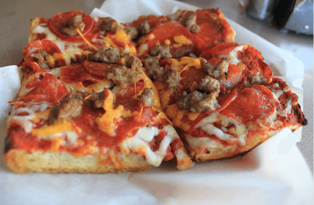 Square slice of pizza with tomato sauce, melted cheese, pepperoni, and sausage on white paper