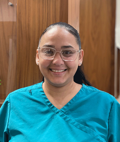 Smiling person in teal scrubs and glasses standing indoors in front of wooden panels.
