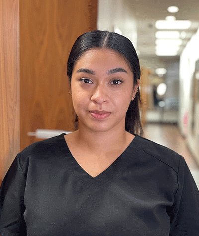 Person in black scrubs standing in a hallway, facing the camera with a slight smile.