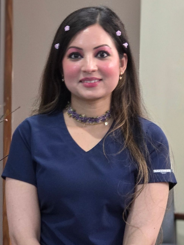 Woman in navy scrubs, standing indoors and smiling, with dark hair and a choker necklace.