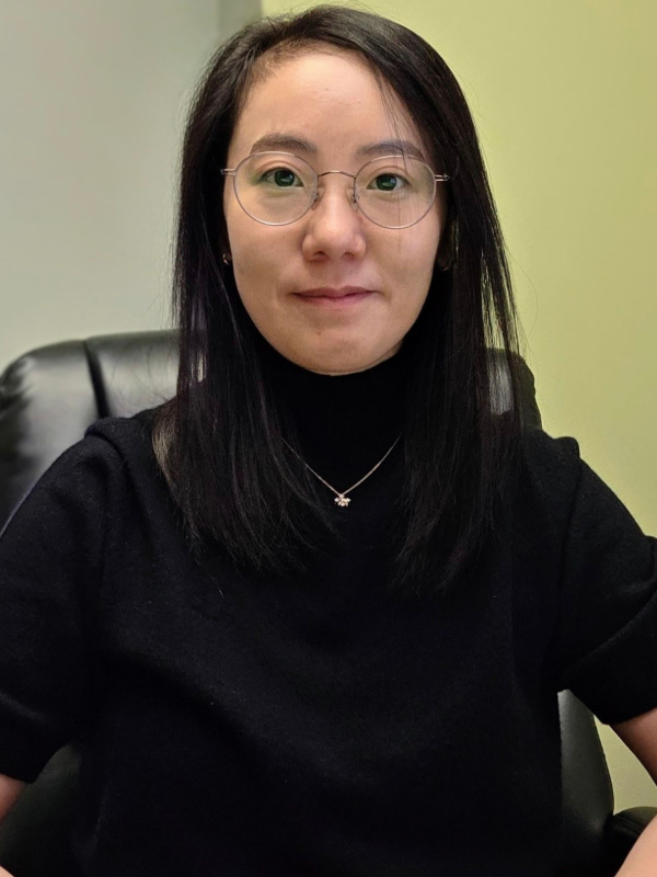 Person with glasses and long dark hair sitting in an office chair, wearing a black top.