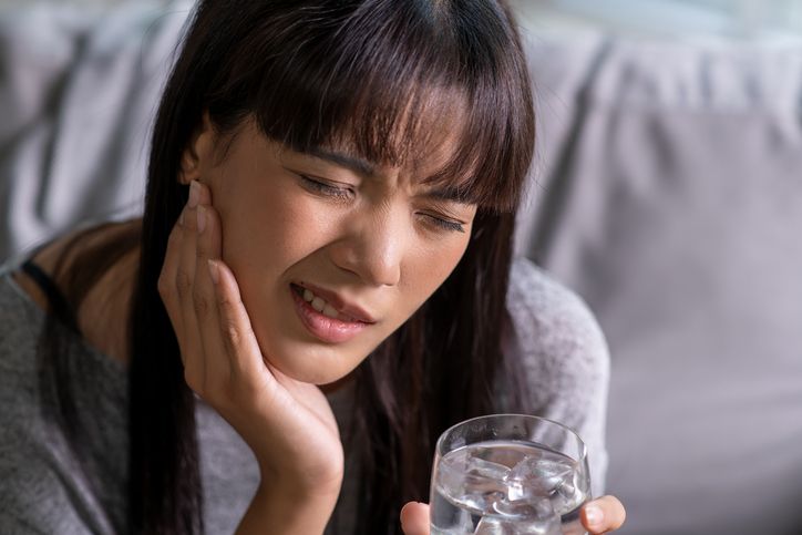 Woman grimacing and holding her cheek while holding a glass of water indoors