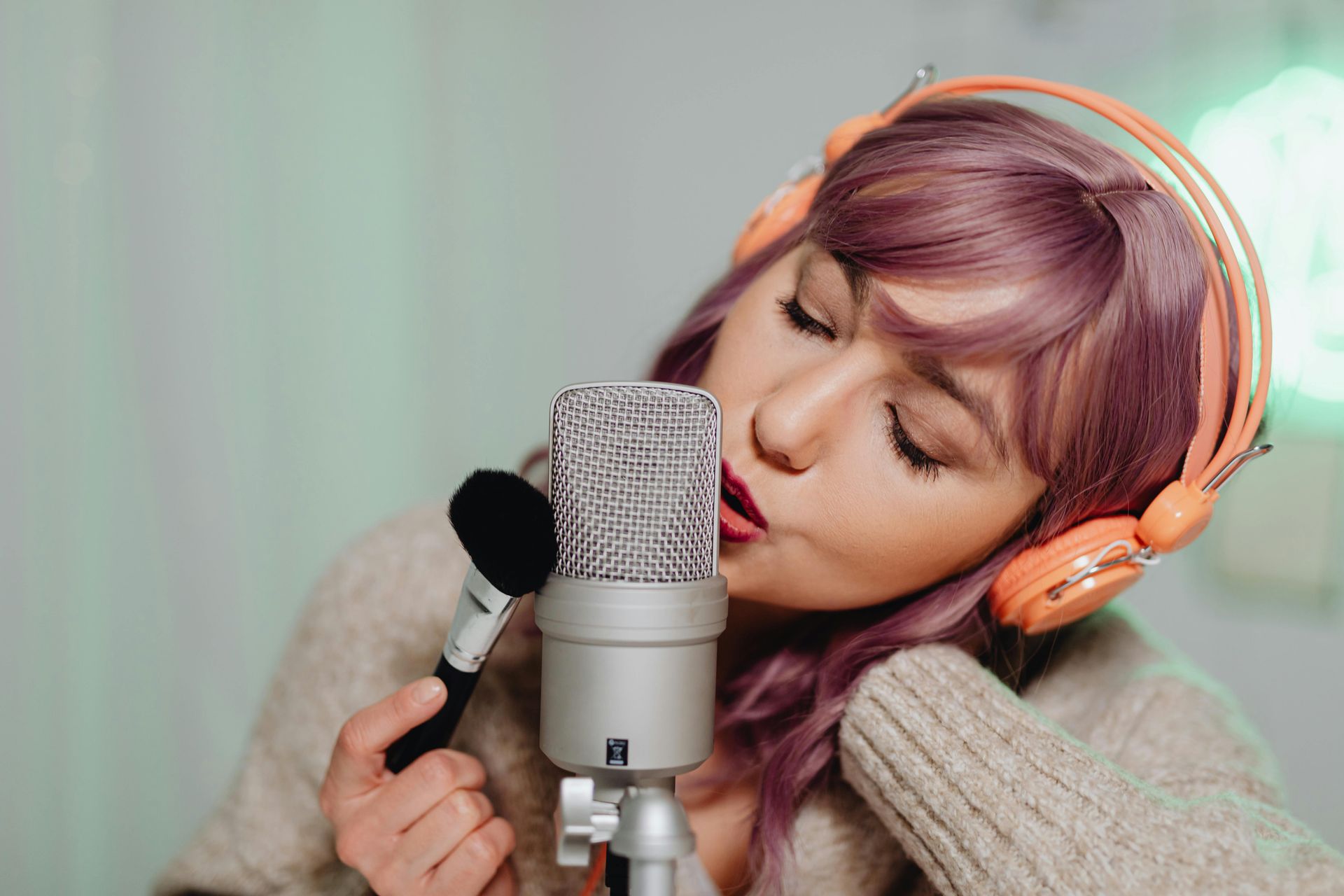 Vrouw met paars haar zingt in een microfoon, draagt ​​een koptelefoon en houdt een make-upborstel vast.