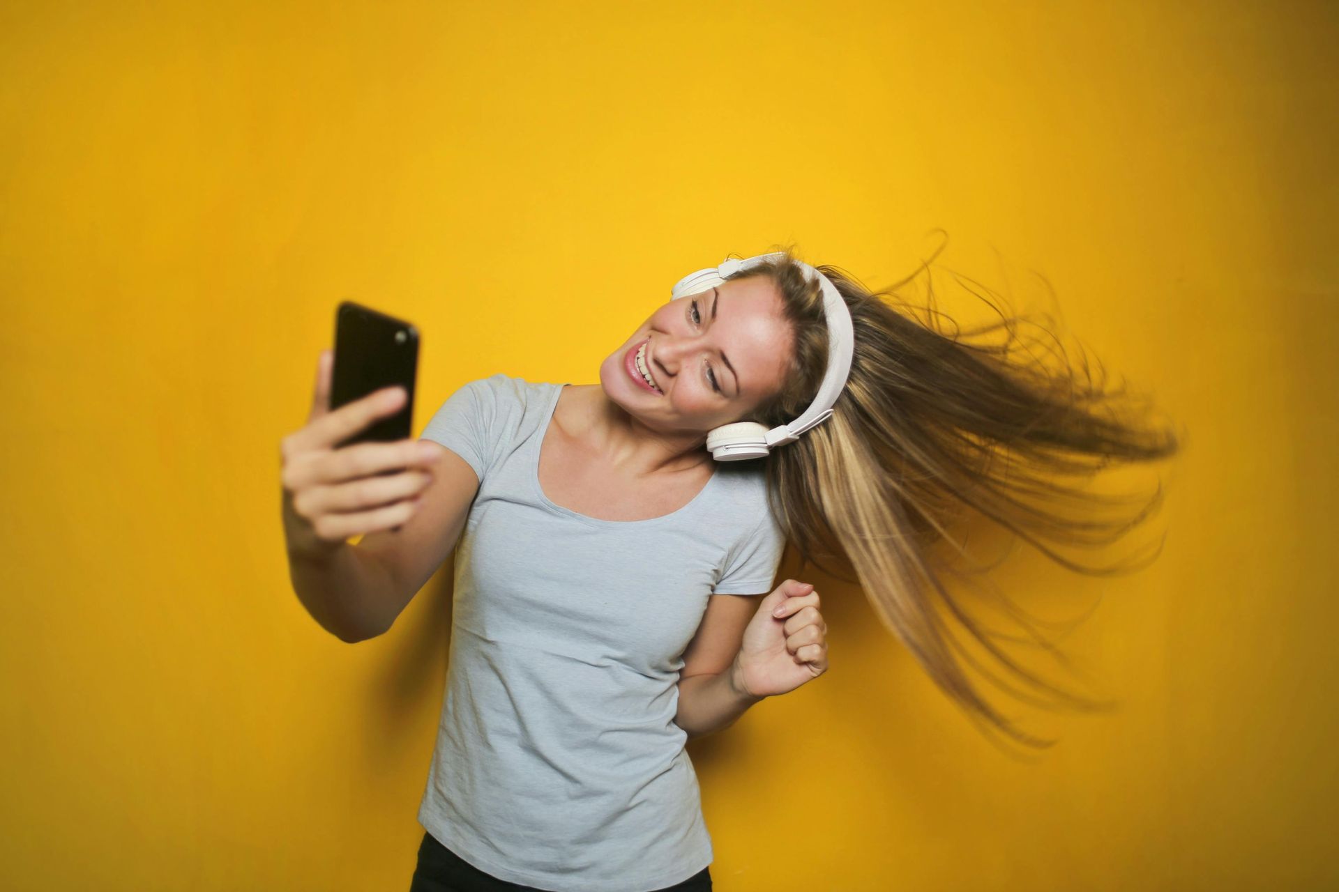 Vrouw met koptelefoon op, telefoon in de hand, danst tegen een gele achtergrond.