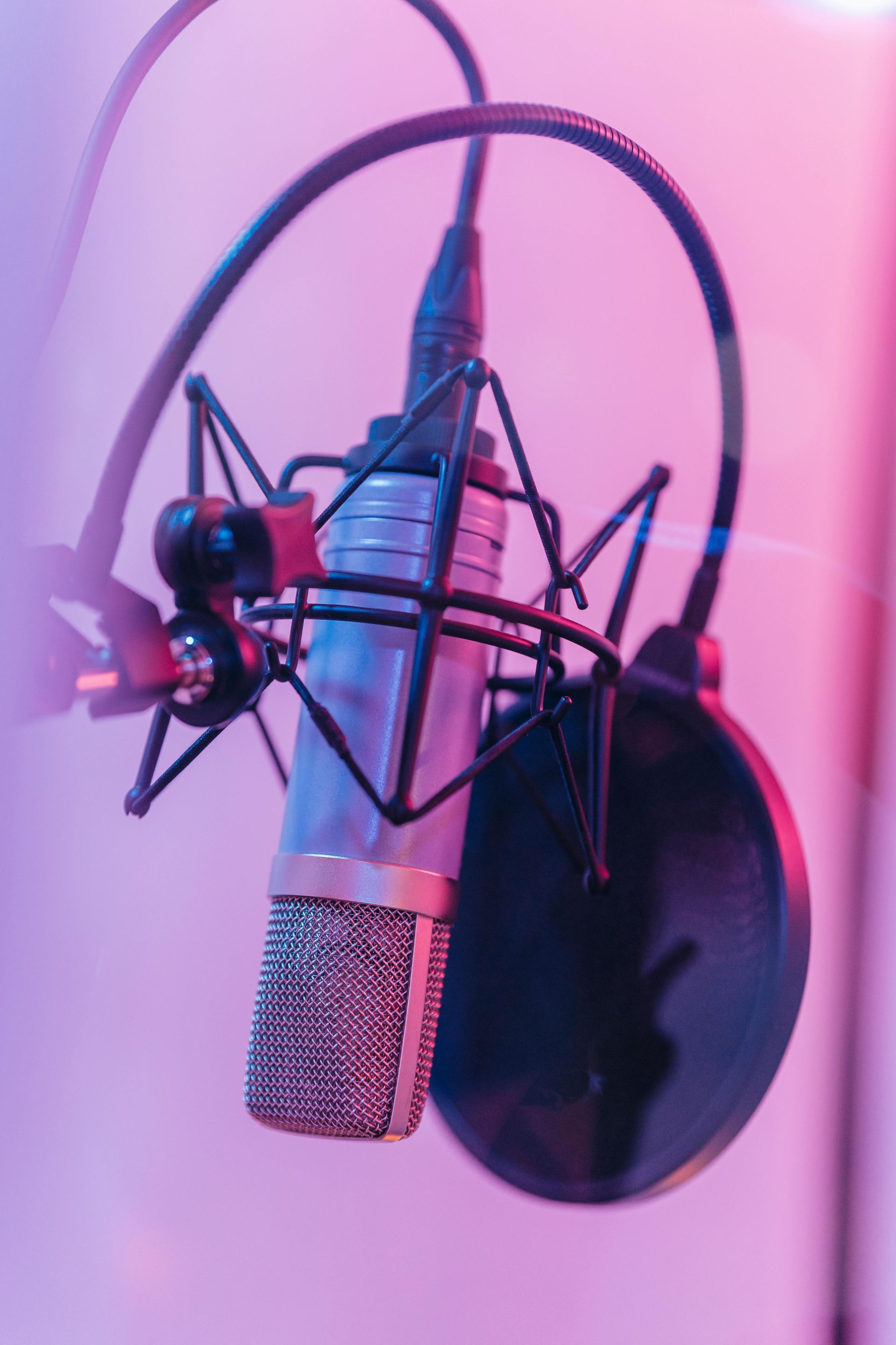 Microfoon in een opnamestudio, opgehangen in een schokdemper en popfilter, badend in roze en blauw licht.