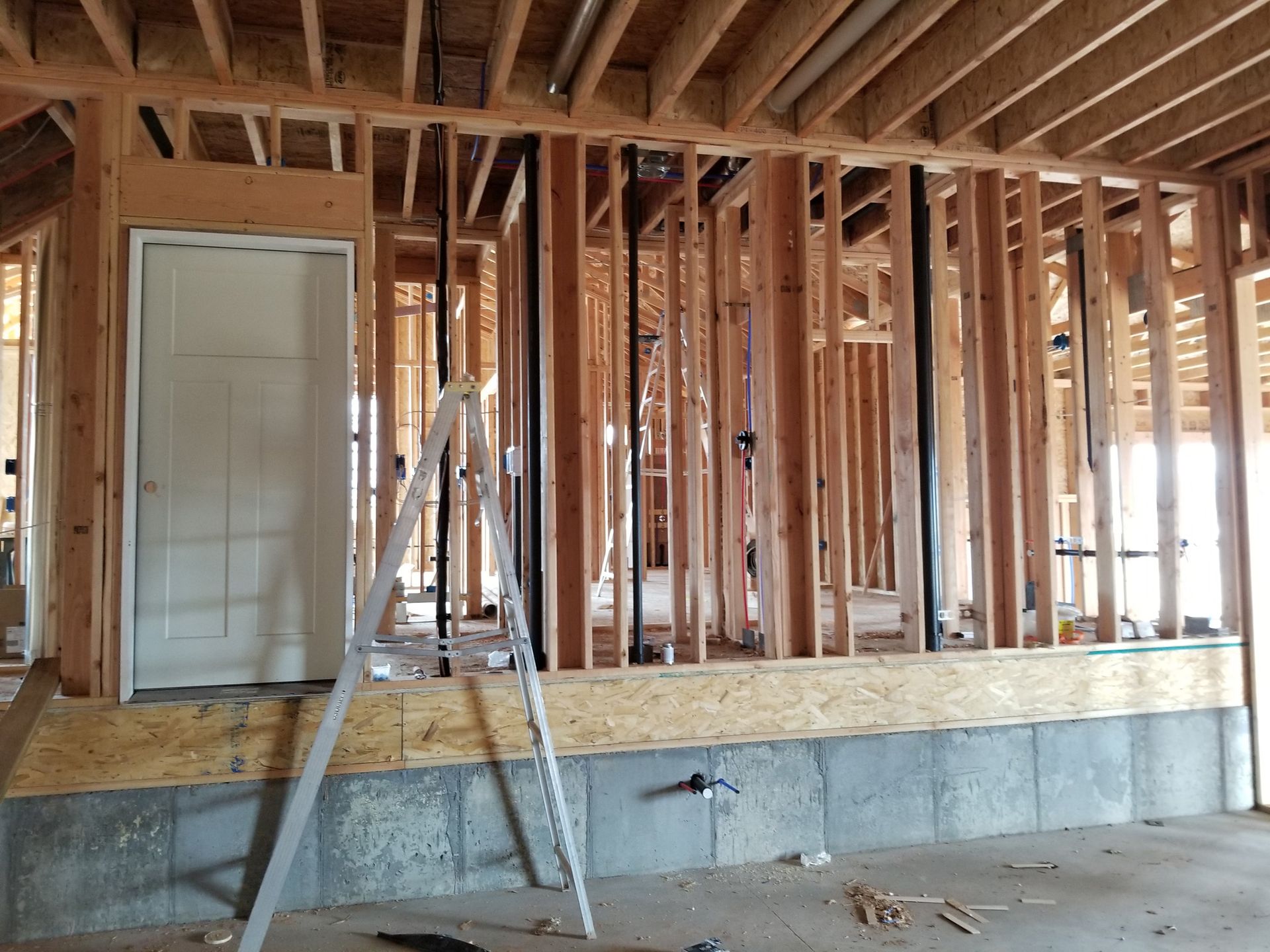 A construction site interior showing wooden wall framing on a concrete foundation, with a door and a ladder in the foreground.