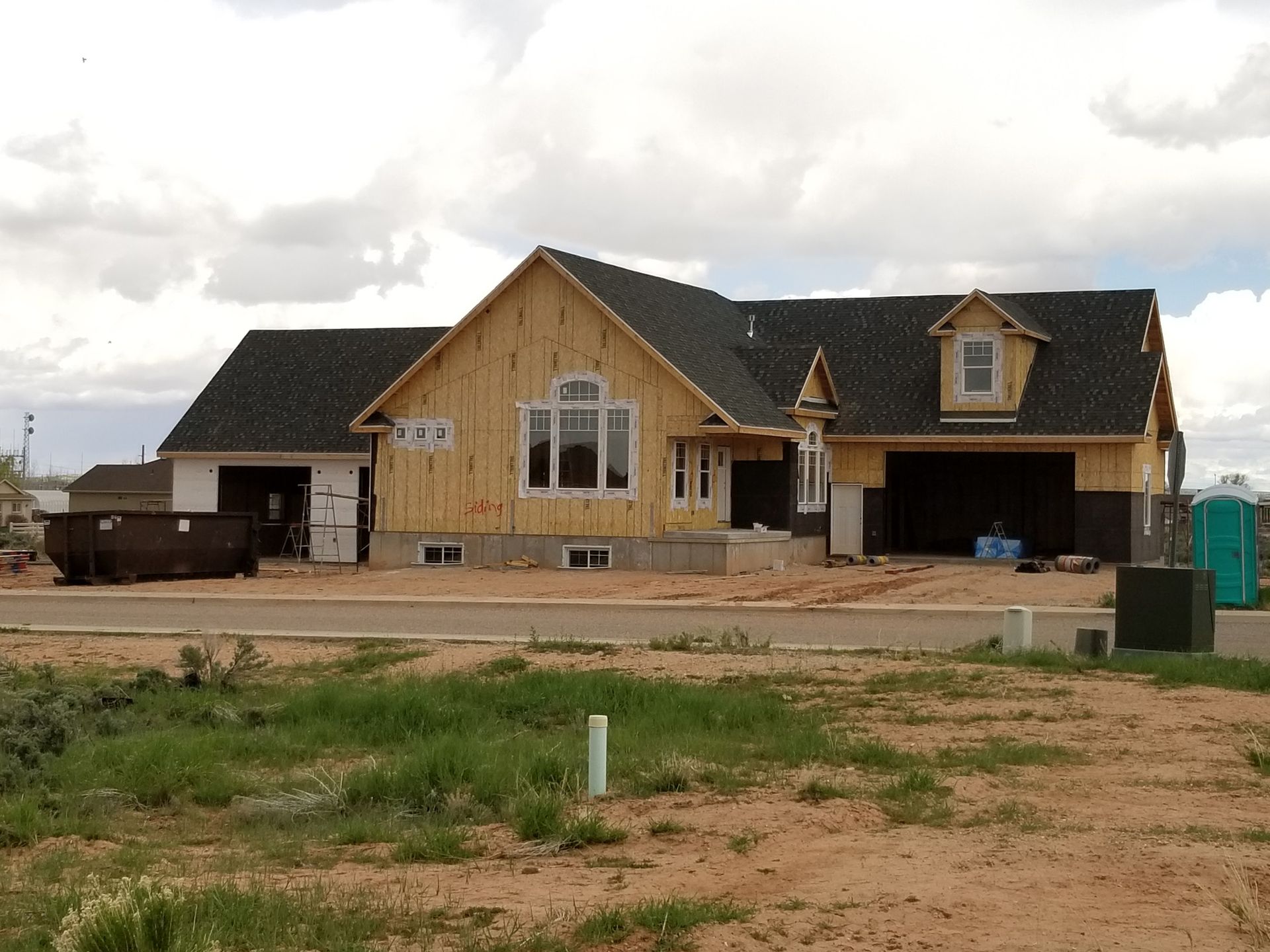 A house under construction with exposed wooden framing, dark roof shingles, and a dumpster in a rural, dirt-lot setting.