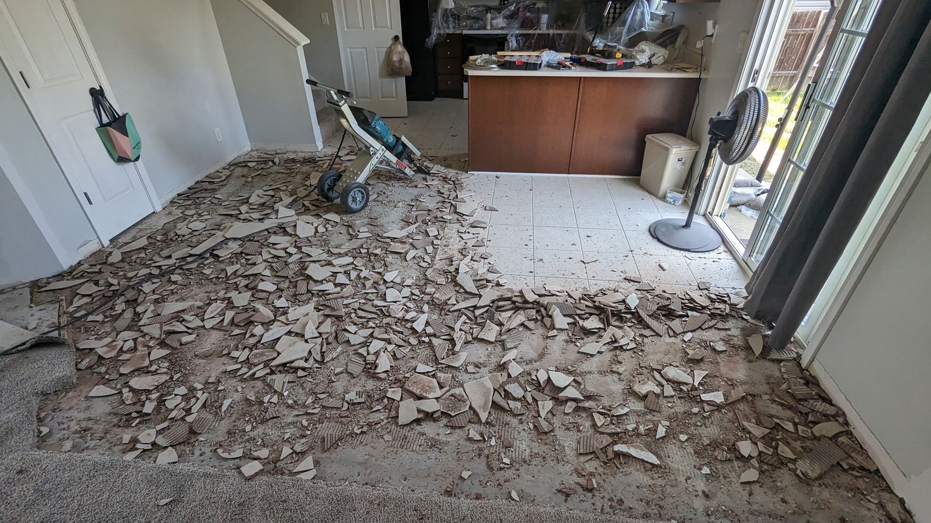 A floor being renovated, with broken tiles and debris covering a section of the room next to a kitchen island.
