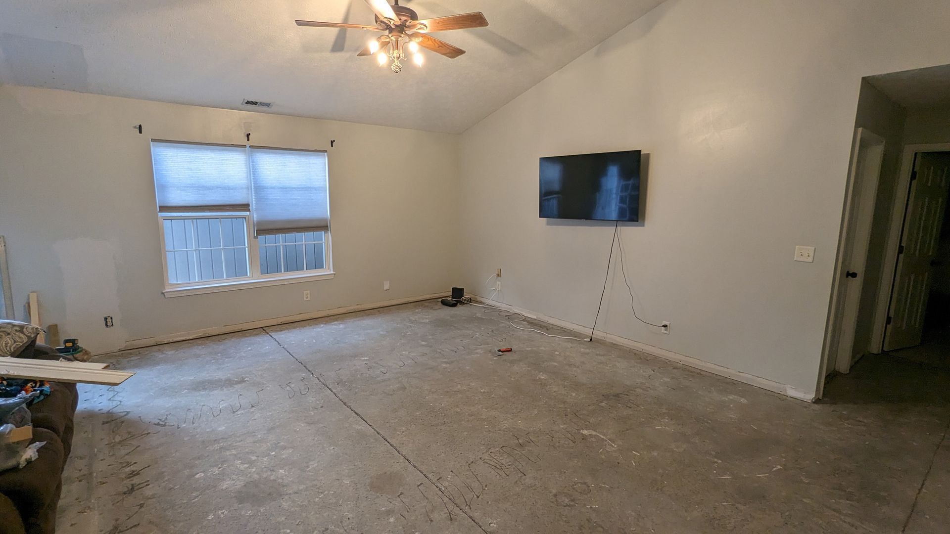 An empty living room with light gray walls, a ceiling fan, a window, and a wall-mounted television over a bare floor.