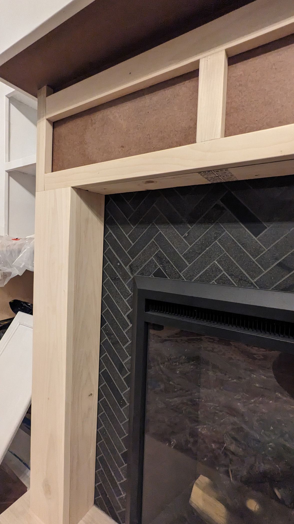 A fireplace under construction, featuring a natural wood frame over dark, herringbone-patterned tile surrounding the firebox.