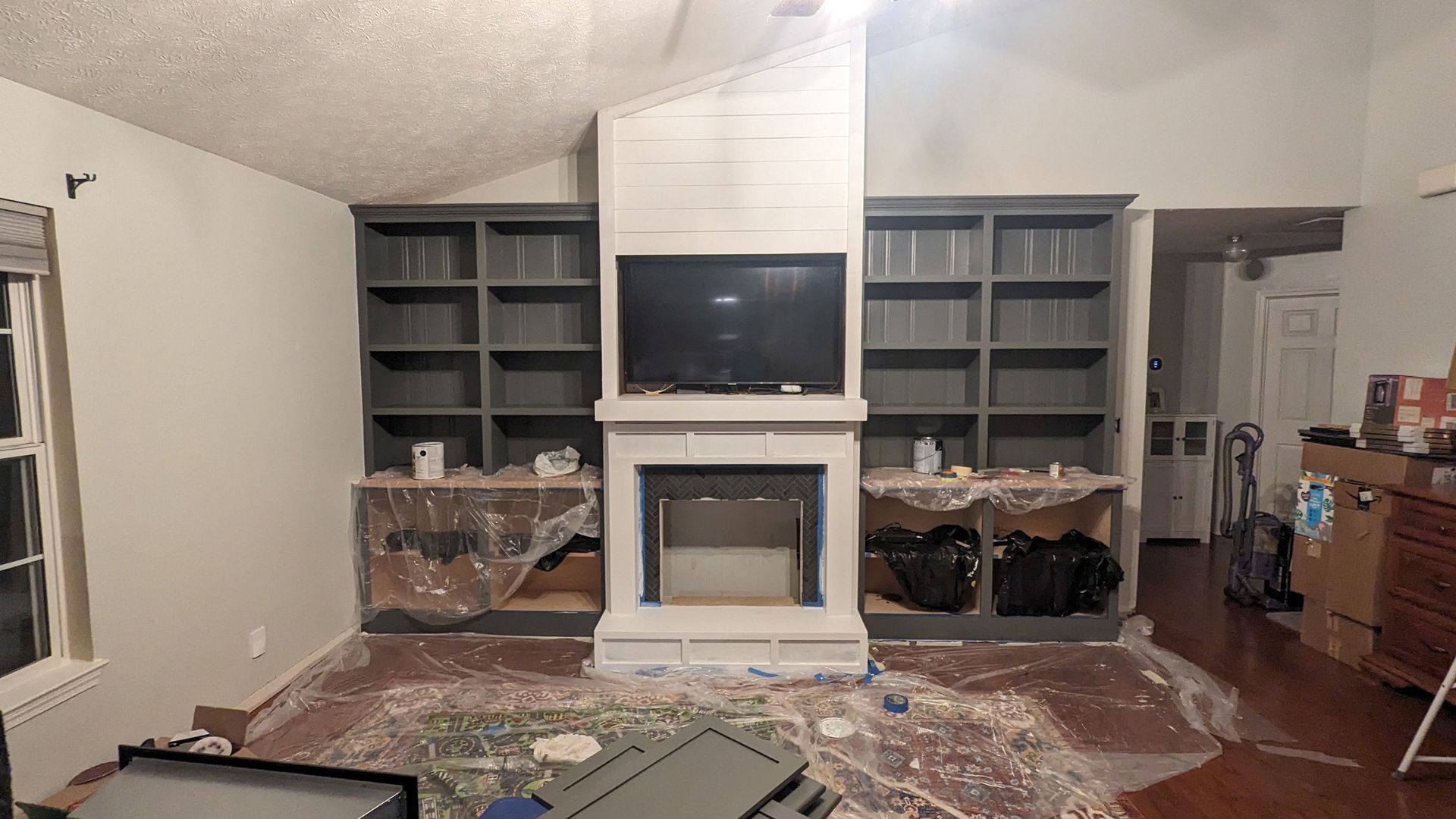 A fireplace unit with a mounted television and dark grey bookshelves, surrounded by plastic drop cloths during a project.