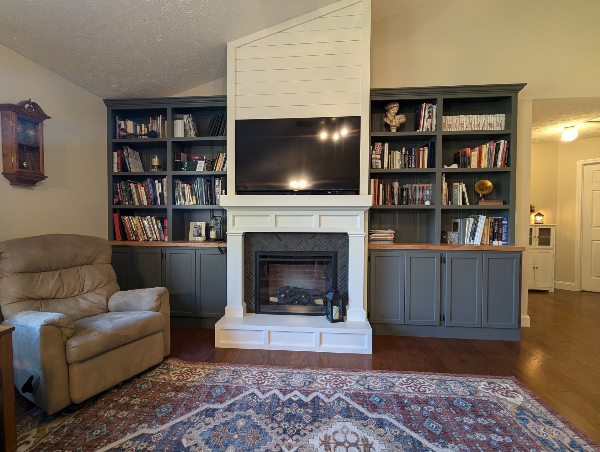A living room with a central fireplace, mounted TV, and dark gray built-in bookshelves on both sides, next to an armchair.
