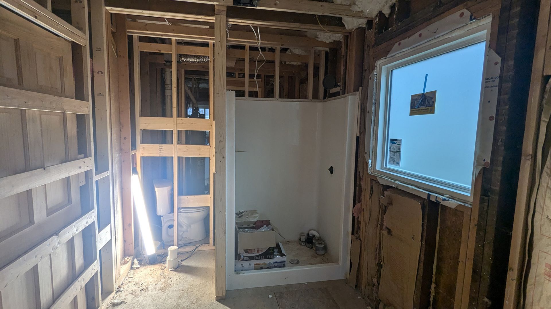 A room under renovation with wooden wall studs, a white shower stall, and a window with a blue sky visible outside.