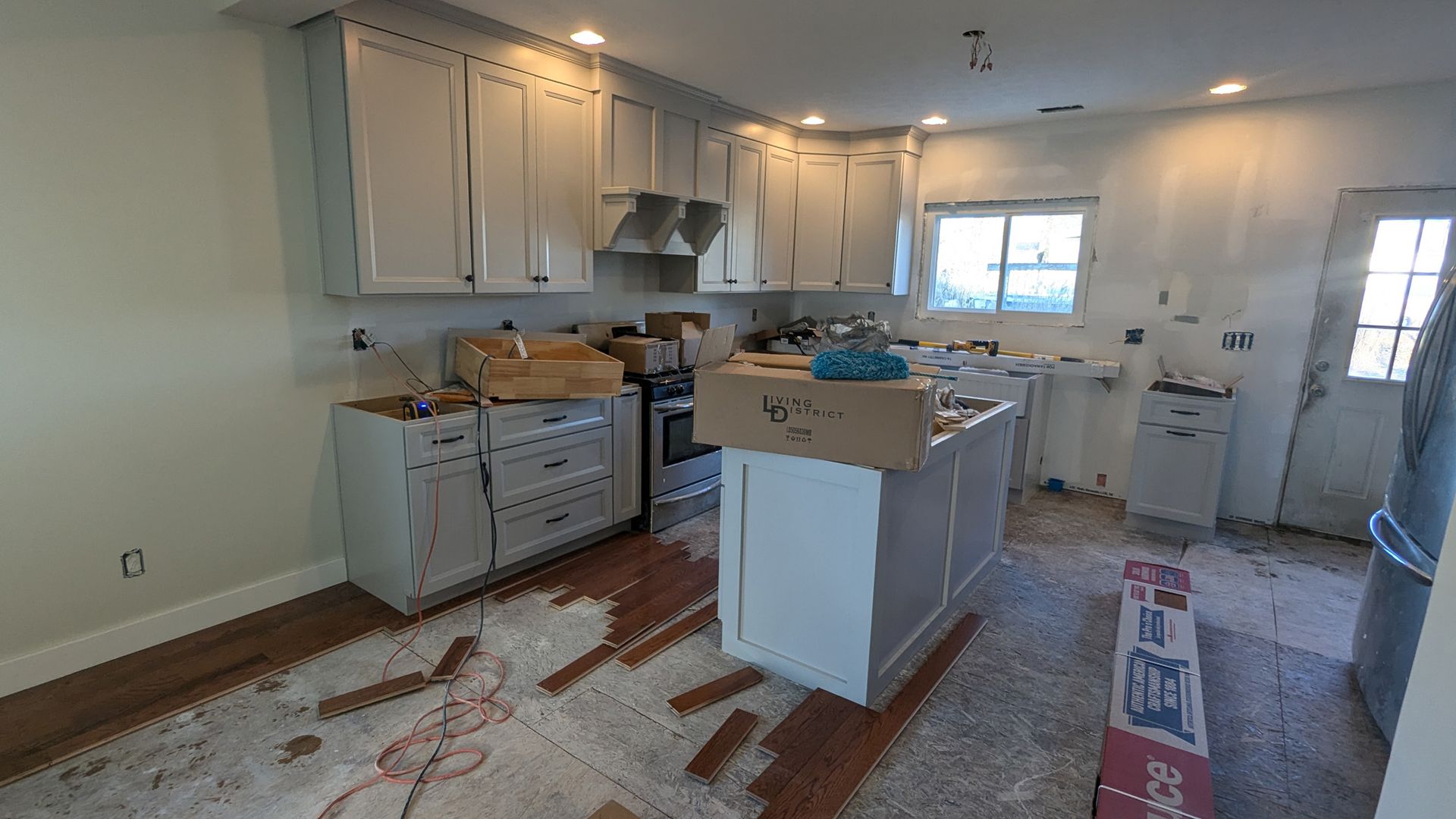 A kitchen under renovation with white cabinets, an unfinished island, wooden floors, and supplies scattered on the floor.