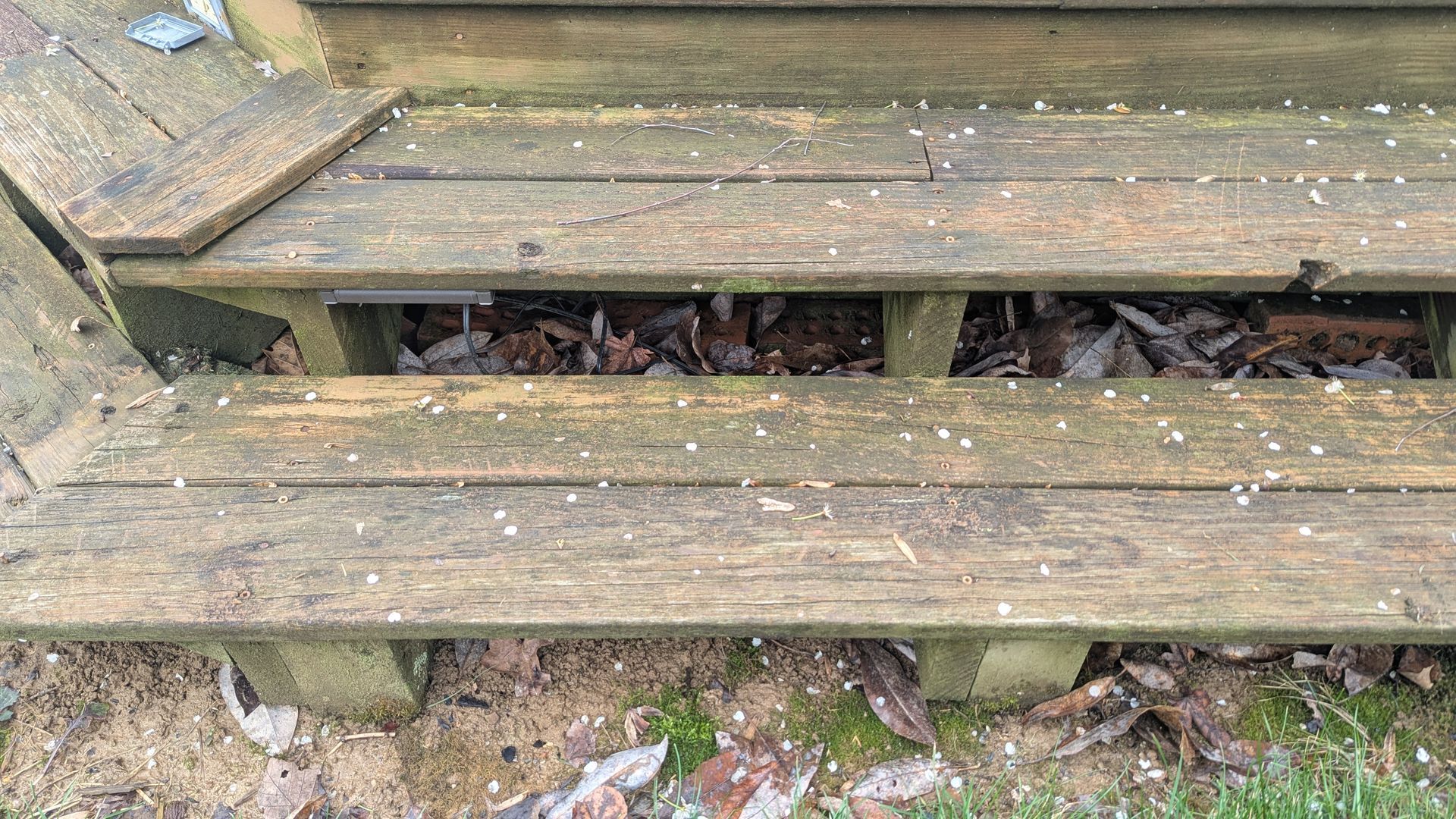 Weathered wooden outdoor steps covered in light scattered snow or hail, with fallen leaves visible underneath.