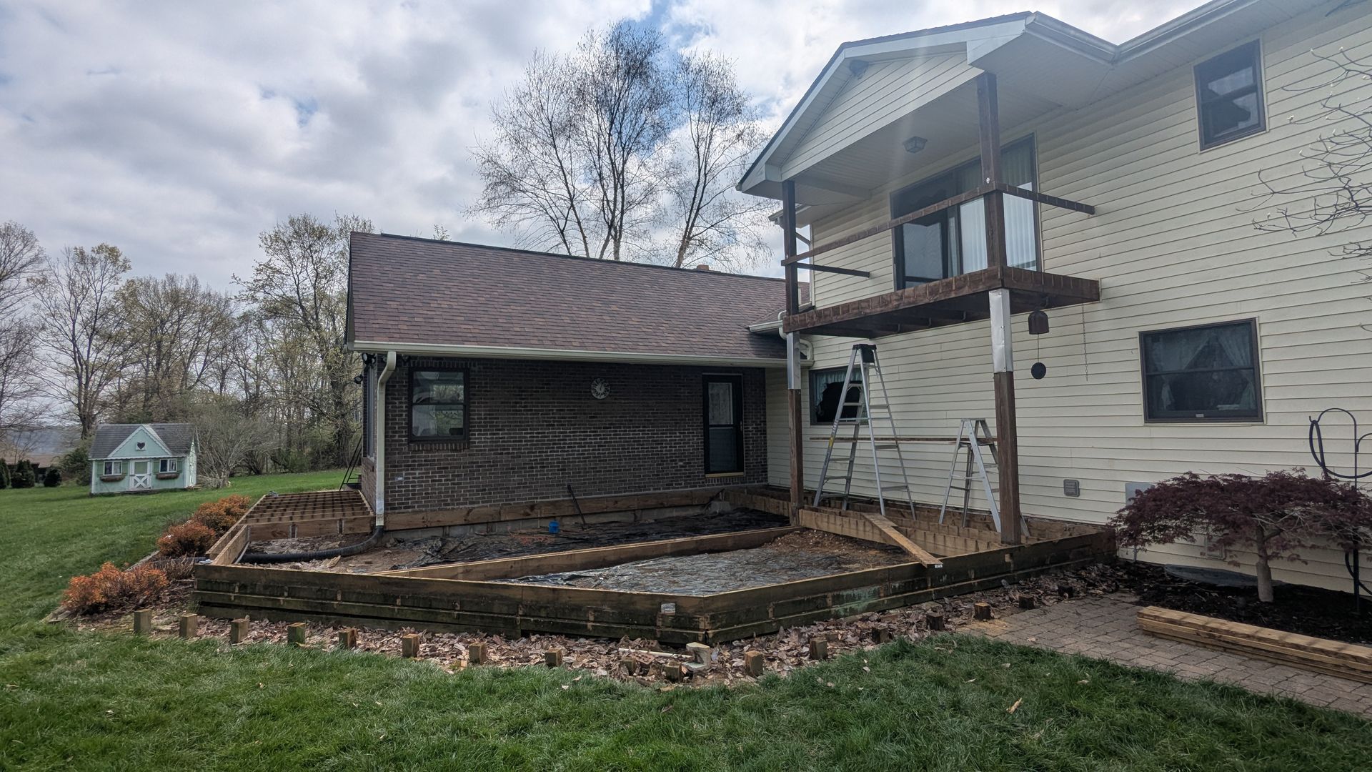 A multi-story house with siding, a wooden deck, and a brown log-style extension, featuring a landscaped patio area.
