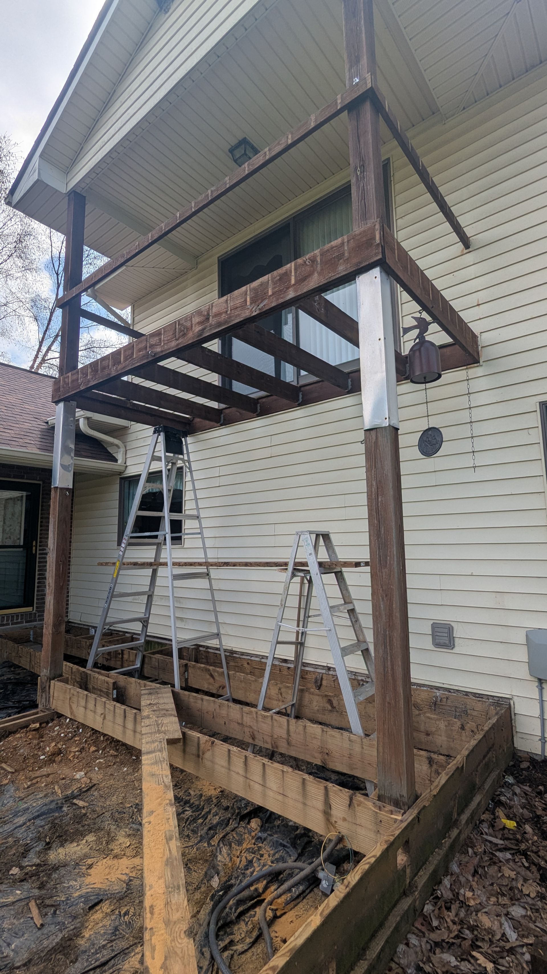 A two-story house deck under construction with exposed wooden framing, support posts, and two ladders placed underneath.