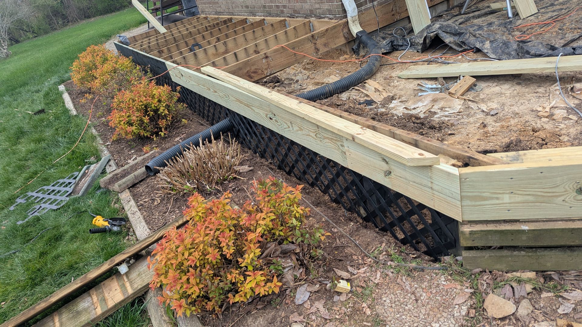 A partially constructed wooden deck with black lattice skirting installed over a garden bed next to a grass lawn.