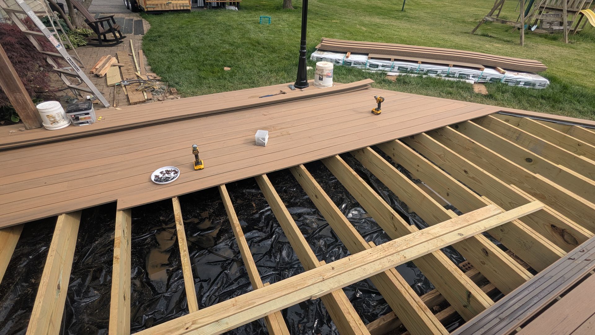 A wooden deck under construction with brown composite planks partially installed over a frame of joists in a grassy yard.