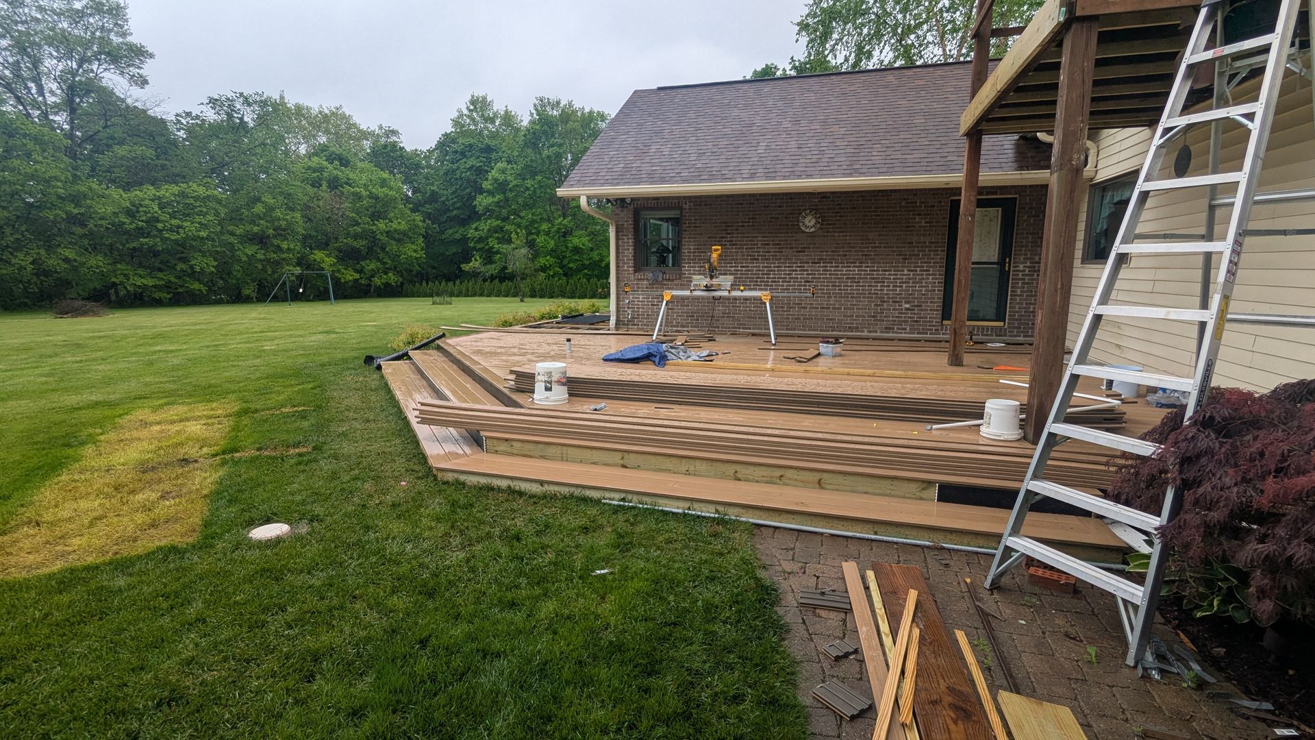 A backyard view of a deck under construction with wood framing and stairs, beside a brick house and a metal ladder.
