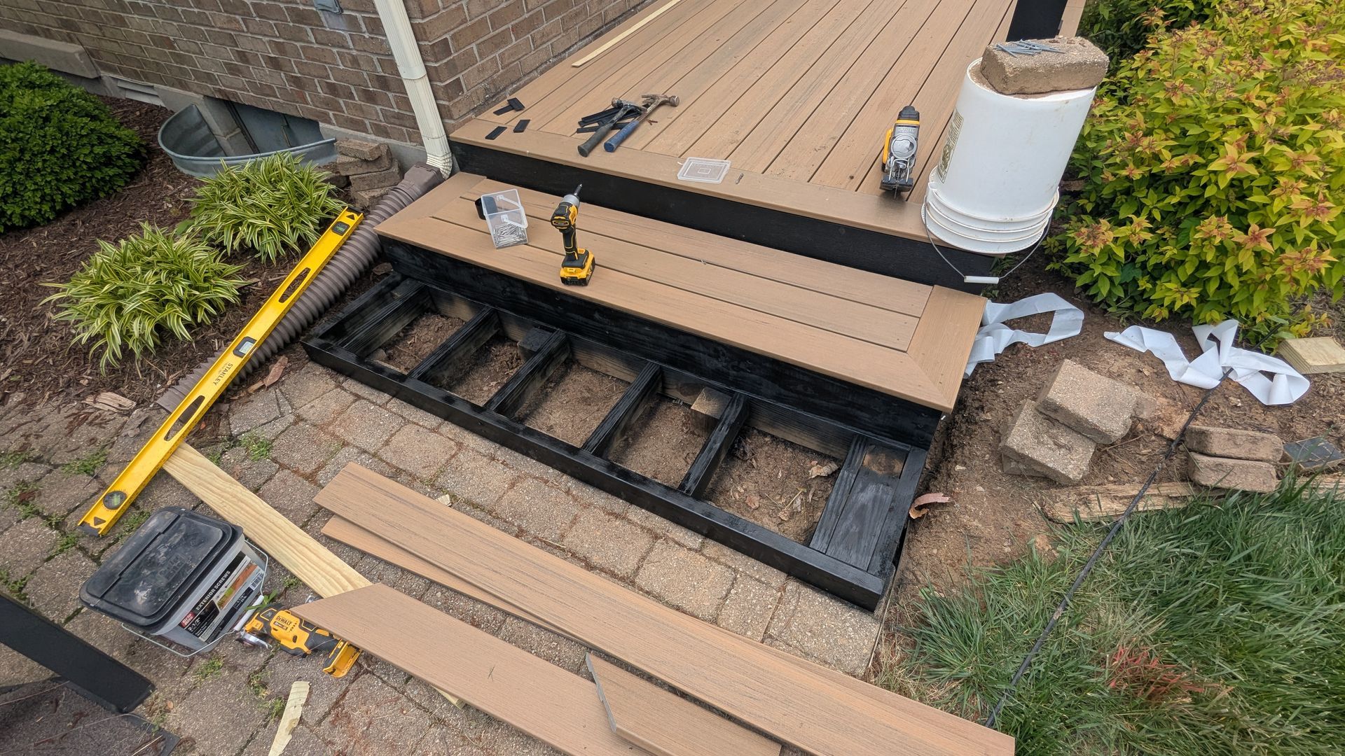 A deck renovation in progress with exposed black framing, tan composite steps, a level, and tools on a paver patio.