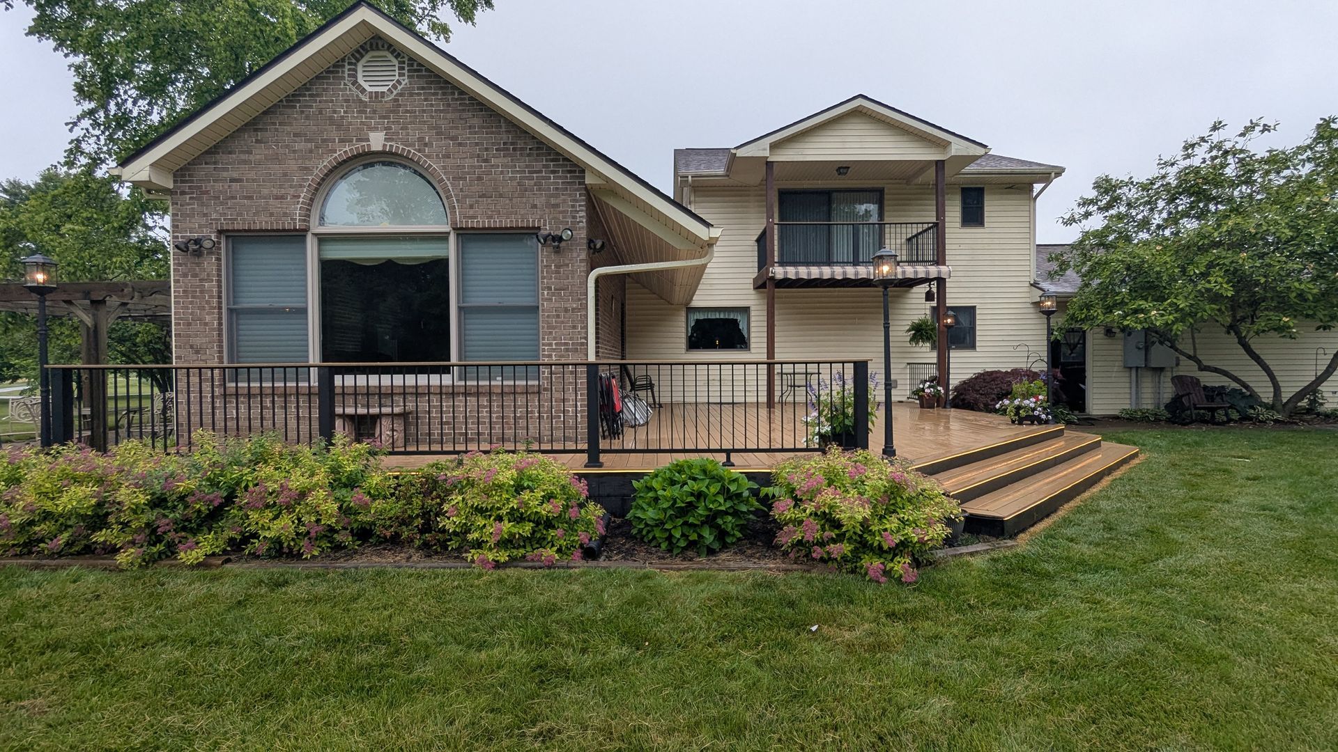 A house with a brick exterior and a cream-colored wing, featuring a large deck with railings and steps in a grassy yard.