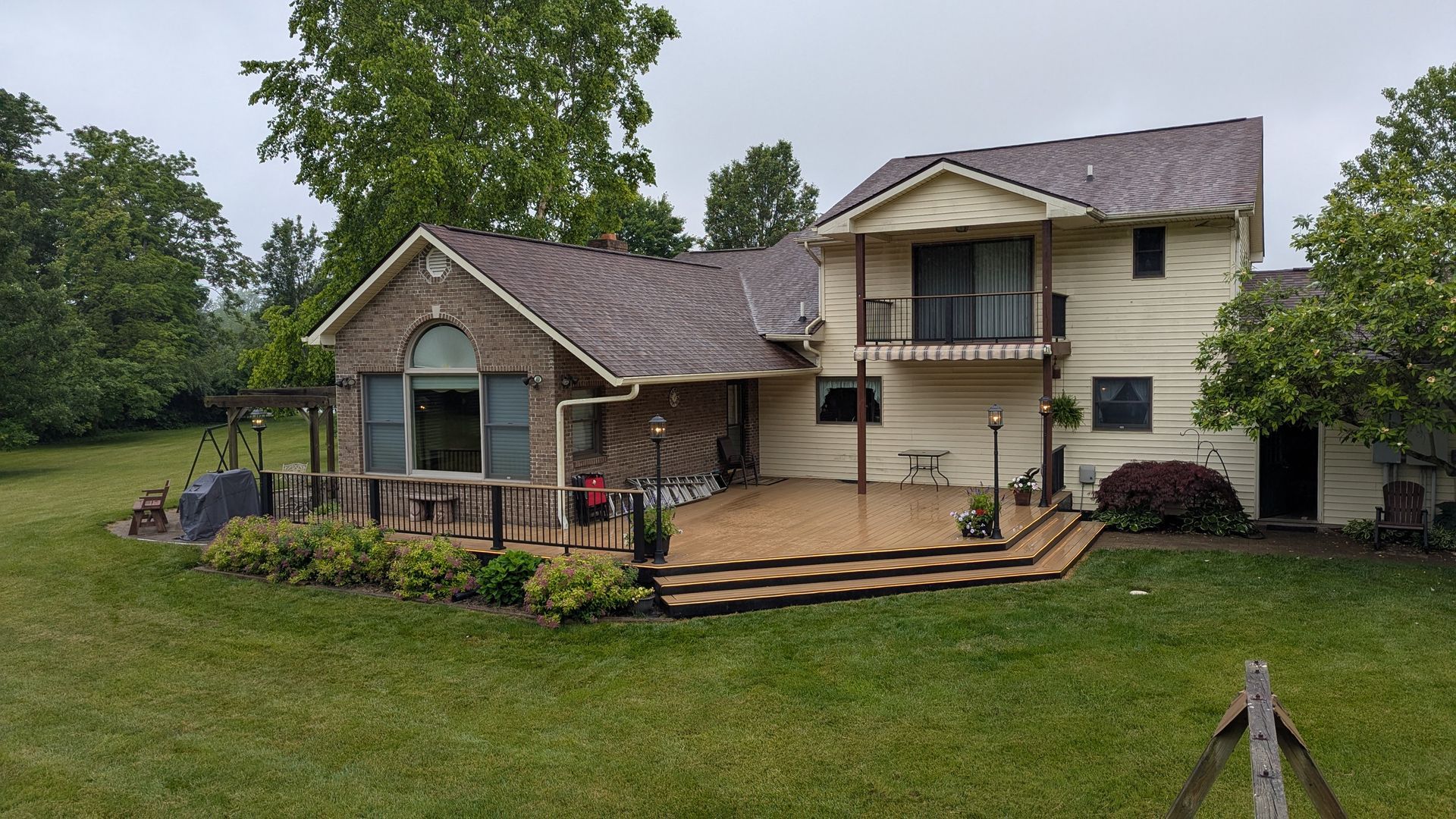 A house with a brick and beige exterior, featuring a large wooden deck and a second-story balcony overlooking a lawn.