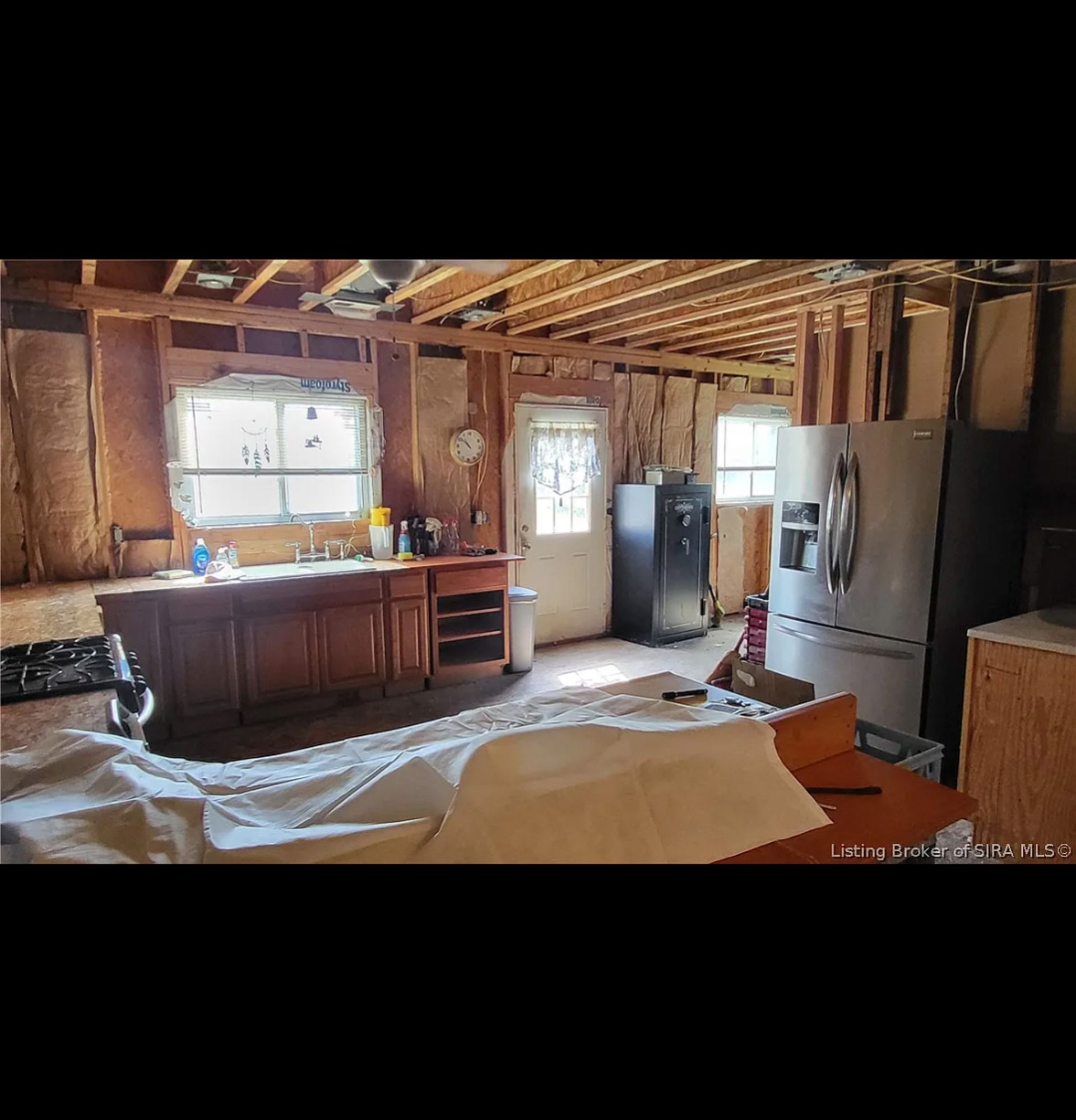 A kitchen under renovation with wood cabinets, a large stainless steel refrigerator, and exposed wall studs.