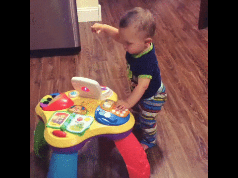 A baby in striped pants plays with a colorful activity table on a wood floor, pointing and pressing buttons.
