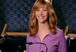 Lisa Kudrow smiling at a microphone in a recording studio. She wears a purple shirt, and has auburn hair.