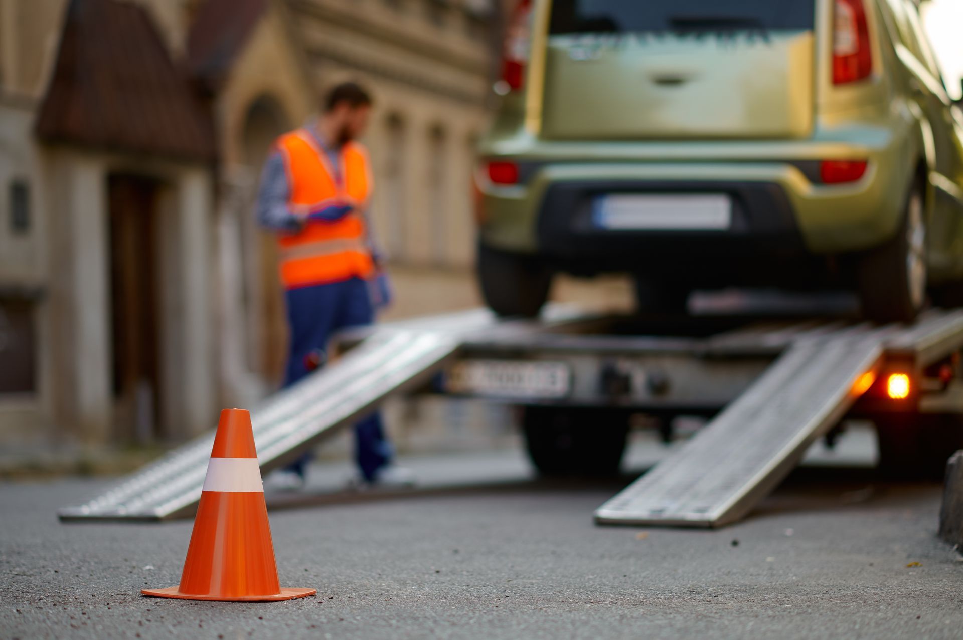 Orange traffic cone in foreground, car being loaded onto a tow truck by a worker wearing a safety vest.