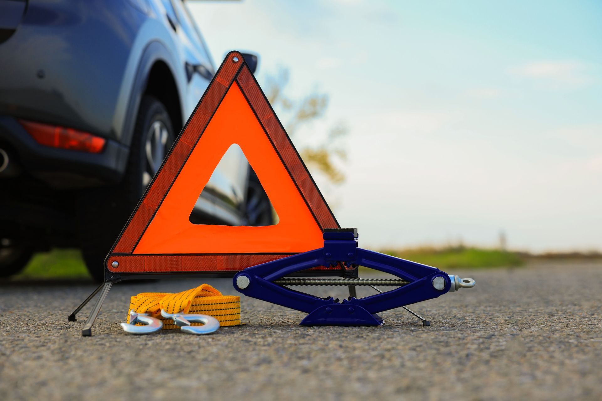Orange safety triangle, jack, and tow rope next to a car on a road.