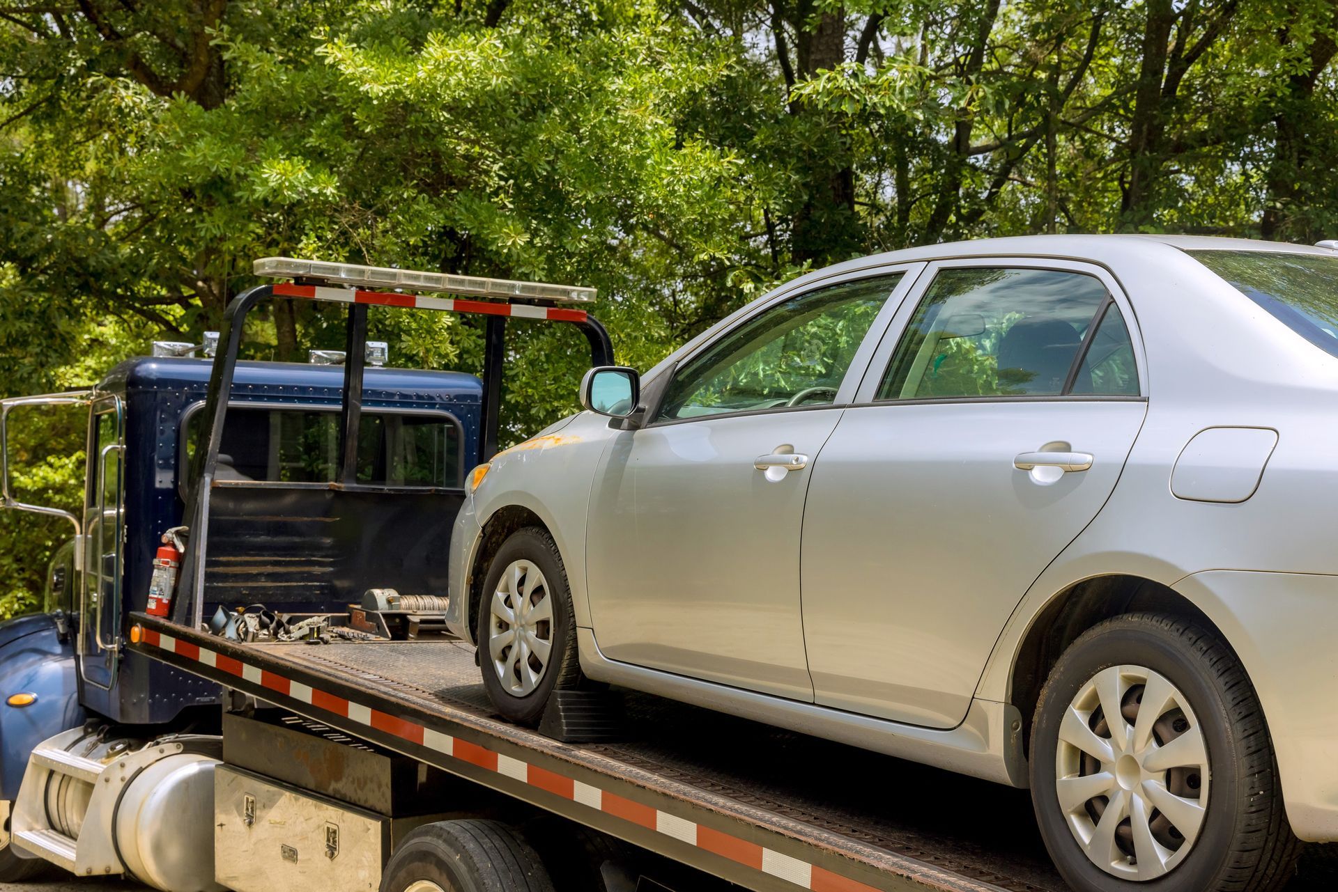 Silver car being towed onto the bed of a tow truck in front of trees.