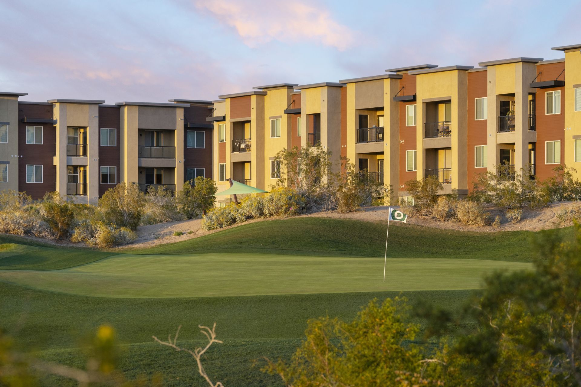 Modern apartment buildings beside a green lawn and landscaped hillside at sunset