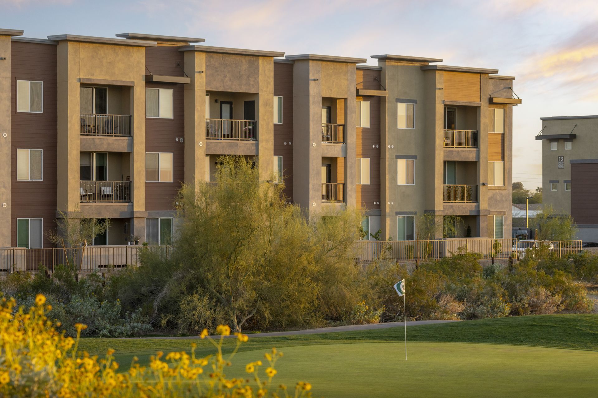 Modern apartment building with balconies, framed by trees and a green lawn at sunset