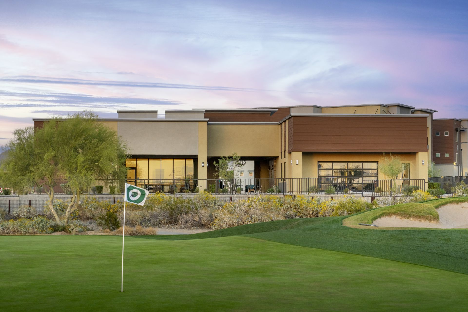 Modern clubhouse overlooking a golf green at sunset, with landscaped shrubs and a flag on the course.