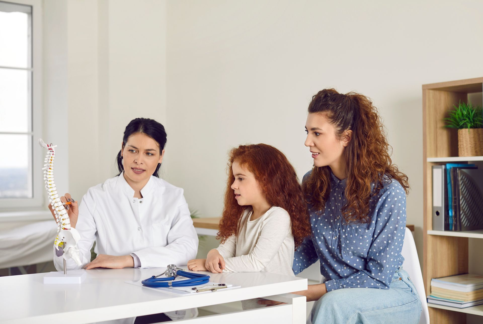 Chiropractor showing a spine model to a young girl and her mother during a family consultation visit