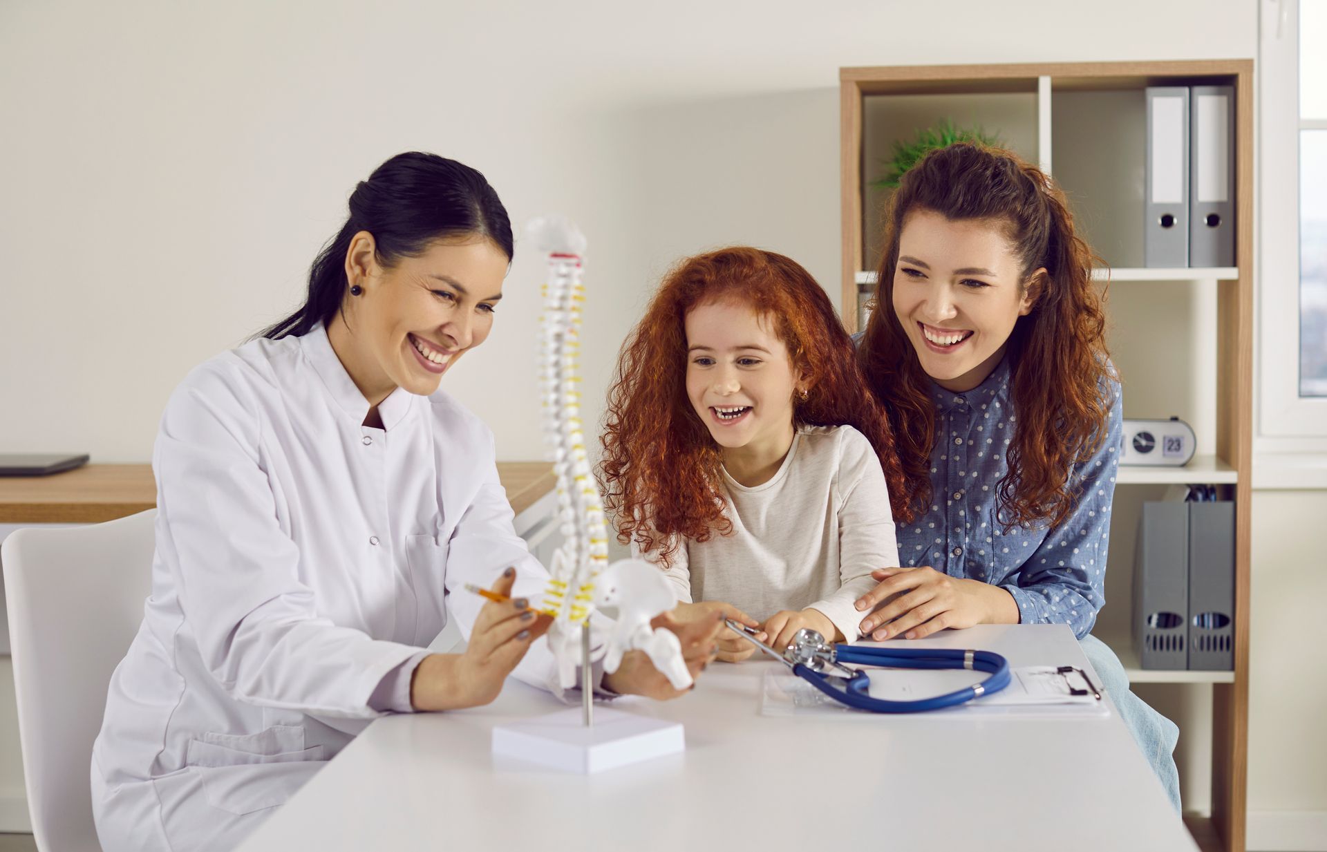 A smiling chiropractor is showing a spine model to a happy young girl and her mother in an exam room A smiling chiropractor is showing a spine model to a happy young girl and her mother in an exam room