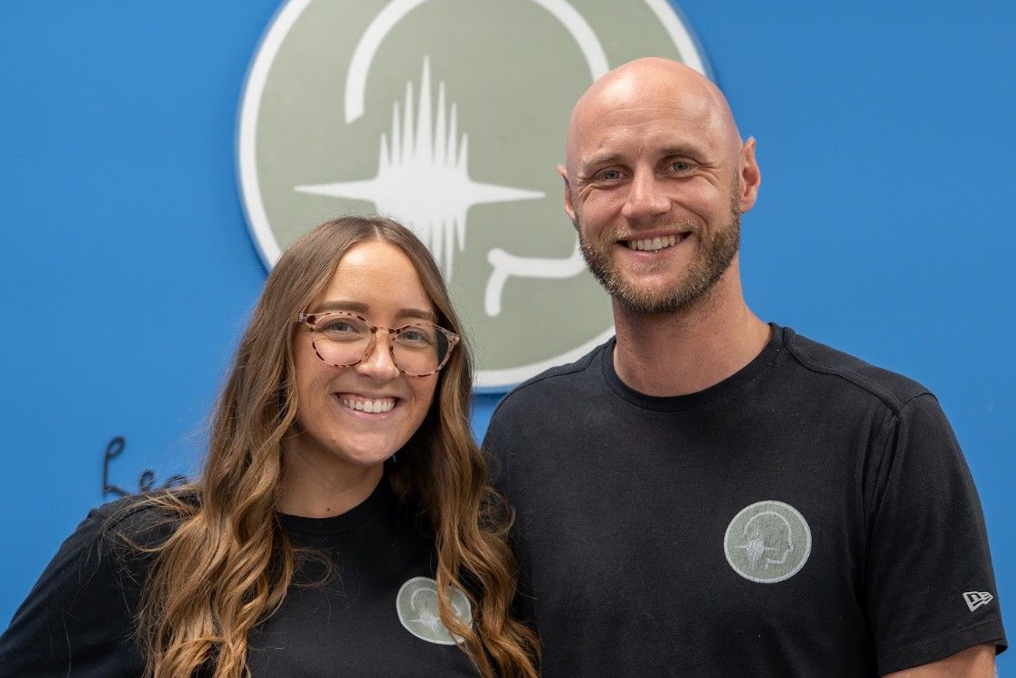 Two smiling people standing before a blue wall with a circular white logo. Two smiling people standing before a blue wall with a circular white logo.