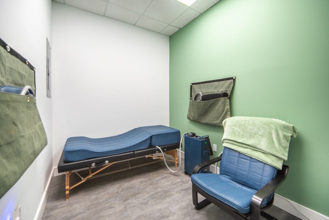 Small exam room with a blue treatment table, blue chair, green walls, and a folded blanket.