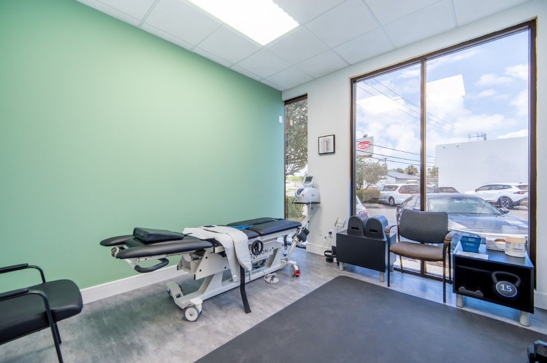 Empty medical exam room with exam table, chairs, and large window.