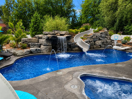 Blue-lit backyard pool at night Backyard pool with rock waterfall, blue water, and a white slide beside a hot tub