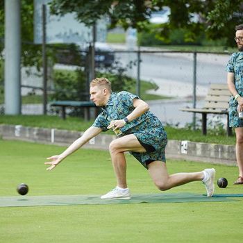 A young male lawn bowler holding a beer and rolling his ball.